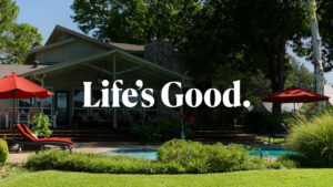 Backyard view of a lakeside home in Granbury, Texas with a pool, red umbrellas, and green landscaping, showcasing a peaceful outdoor living space with the phrase 'Life’s Good.' displayed across the image.